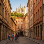 Quatre personnes marchant sur une rue pavée typique de Lyon, encadrée par des immeubles ocres. La basilique de Fourvière domine le paysage.