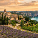 Village perché provençal avec église et maisons en pierre, entouré de champs de lavande et vignes, au-dessus d'une rivière turquoise, sous un ciel orangé.