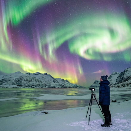 Comment voir les aurores boréales et réussir ton voyage Un photographe en tenue d'hiver capture des aurores boréales vertes et violettes au-dessus d'un lac gelé et de montagnes enneigées.