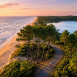 Vue aérienne d'une côte au coucher du soleil. Plage, océan, pinède, lac et piste cyclable avec cycliste. Un surfeur marche sur la plage.