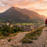 Deux randonneurs contemplent la ville de Bonneville, son château et les montagnes alpines baignées par un coucher de soleil.