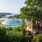 Vue aérienne du Domaine des Ormes avec une cabane perchée, un dôme aquatique, un lac et la mer en arrière-plan.