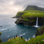 Puffins sur des falaises verdoyantes des îles Féroé, face à l'océan. La cascade de Múlafossur s'y jette. Ciel nuageux.
