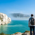 Un randonneur face à la mer et aux falaises du Nord-Pas-de-Calais, avec des bâtiments modernes et anciens en arrière-plan.