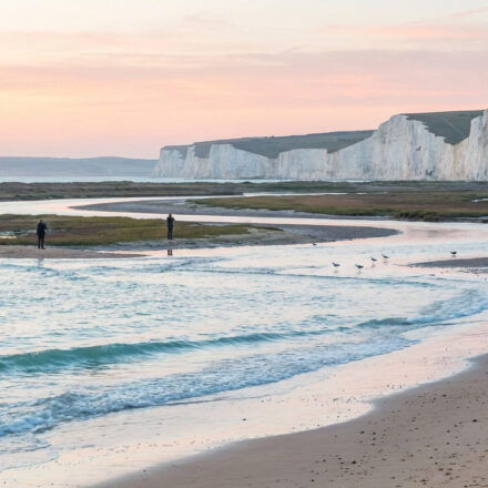 S’évader pour un week-end dans les Hauts-de-France Paysage côtier avec falaises de craie blanches, un estuaire sinueux, une plage de sable et des vagues sous un ciel rose pâle. Deux silhouettes lointaines.
