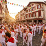 Vue d'une rue bondée à Bayonne, des milliers de personnes en tenue blanche et foulard rouge célèbrent sous des guirlandes colorées.