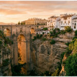 Vue panoramique du Pont Neuf de Ronda traversant une gorge escarpée, avec des bâtiments blancs perchés sur les falaises sous un ciel orangé.