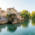 Vue panoramique de l'Isle-sur-la-Sorgue avec son canal turquoise, une grande roue à aubes en bois, des bâtiments en pierre et des saules pleureurs le long des berges animées.