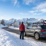 SUV garé sur route enneigée avec skis sur le toit, entouré de montagnes et forêts. Deux personnes préparent l'équipement.