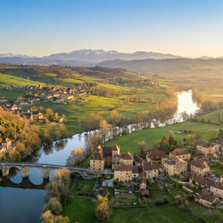 Vue aérienne de Carennac (Lot) : château médiéval, village pittoresque, rivière sinueuse et pont en pierre, avec montagnes au loin.