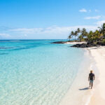 Un homme marche sur une plage de sable blanc bordée de palmiers, face à une mer turquoise et limpide à l'île Maurice.