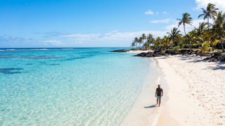 Un homme marche sur une plage de sable blanc bordée de palmiers, face à une mer turquoise et limpide à l'île Maurice.
