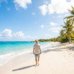 Vue arrière d'un homme en chapeau marchant pieds nus sur une plage tropicale, mer turquoise et palmiers luxuriants.