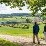 Un couple de randonneurs sur un sentier en campagne, un train Transilien traversant une vallée verdoyante sous un ciel nuageux.