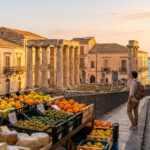 Un homme sur une rue pavée de Syracuse, contemplant la mer. Au premier plan, un marché coloré. Au fond, des ruines antiques.