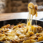 Une fourchette soulève une pomme de terre d'une truffade fumante, montrant le fromage fondu et filant dans une poêle en fonte.