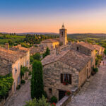 Vue aérienne d'un village provençal en pierre avec des toits de tuiles, une église, des ruelles fleuries et des vignobles au loin au crépuscule.