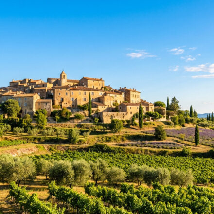Que faire en Provence : les plus beaux endroits à découvrir Un village provençal perché, en pierre ocre, avec son clocher, dominant des champs de vignes verts et de lavande violette sous un ciel bleu clair.