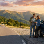 Couple planifiant un road trip sur tablette, près d'un SUV bleu, sur une route de montagne sinueuse au lever/coucher du soleil.
