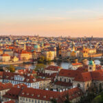 Panorama de Prague au crépuscule. La Vltava serpente entre les bâtiments aux toits rouges, dominés par l'Église du Týn et le Pont Charles.